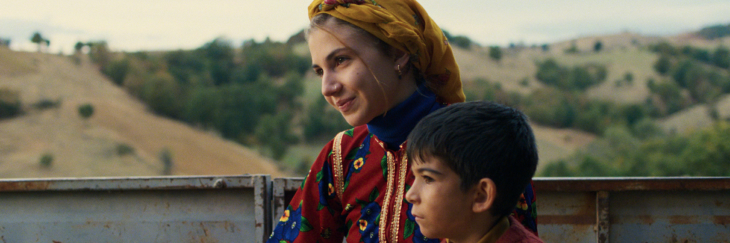 Une jeune fille habillée en vêtements traditionnels colorés et un enfant sont assis.es à l'arrière d'un tracteur. 