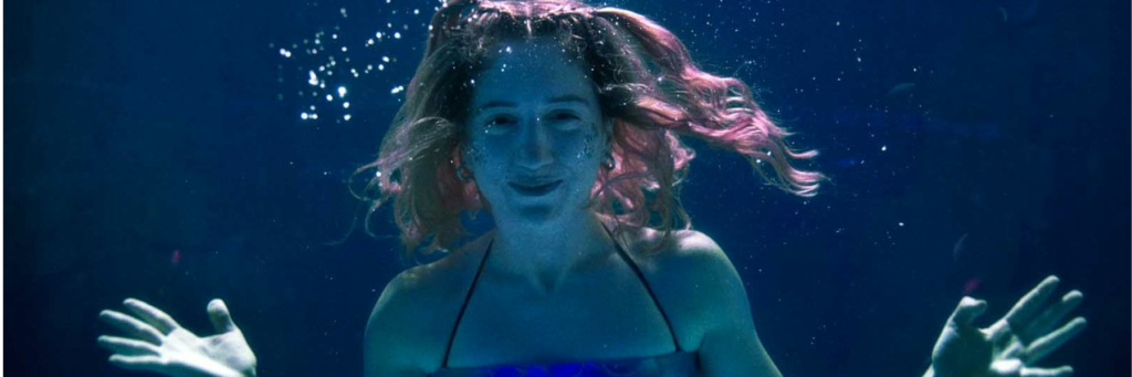 Une jeune femme est dans l'eau, les cheveux roses et elle regarde la caméra. 