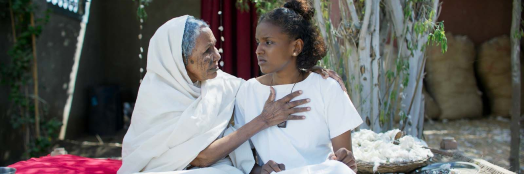 Deux femmes en blanc côté à côté. La grand-mère a la main posée au niveau du coeur de sa petite fille. Elles se regardent dans les yeux.